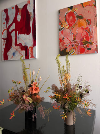 Two abstract paintings on a wall with two floral arrangements on a table below.