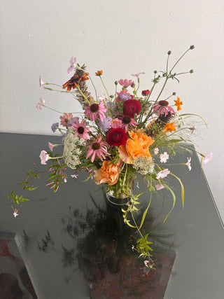 A wildflower bouquet with orange, red, pink, and white blossoms and green foliage, arranged against a white wall and a dark reflective surface. For delivery in los angeles