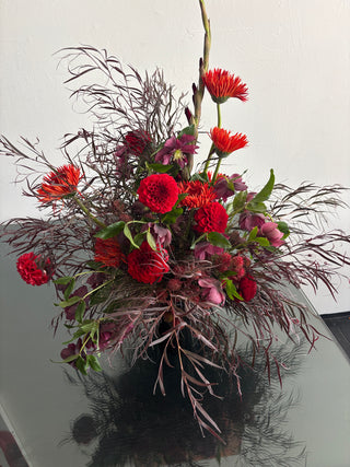 A vibrant floral arrangement featuring bright red gerbera daisies, deep burgundy roses, and dark burgundy feather grass, set in a black vase on a reflective glass table against a plain white wall.