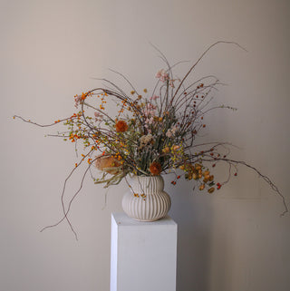 Dried floral arrangement in a textured vase on a white pedestal against a plain wall.