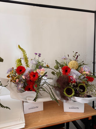 Two wrapped flower bouquets arranged diagonally on a light-colored wooden table against a white backdrop. The bouquets feature prominent red gerbera daisies, green sunflowers, purple wildflowers, and green foliage, wrapped in white paper with small white cards attached.