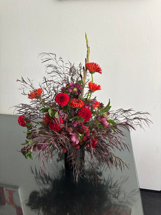 A vibrant floral arrangement featuring red and orange daisy-like flowers, deep pink alstroemeria, and feathery burgundy foliage, presented in a dark vase on a reflective dark surface against a plain white wall.