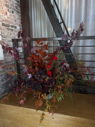 An autumnal floral arrangement in a ceramic vase, featuring burgundy, red, orange, and purple foliage with small berries, displayed on a wooden floor in front of a window and staircase.