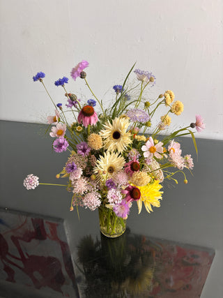 A wildflower bouquet in a small clear glass vase, featuring a mix of purple, yellow, pink, and white flowers, displayed on a dark reflective surface against a plain light gray wall.