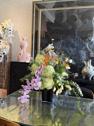 Floral arrangement on a glass table with decorative vases and sculptures in the background.