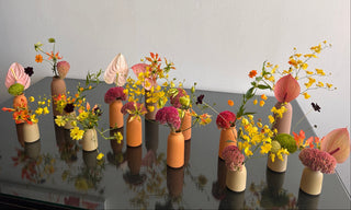 Row of small floral arrangements in various vases on a reflective surface.
